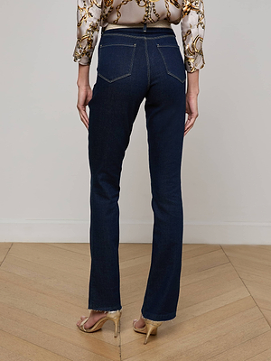 A person stands on a wooden floor, facing away, wearing L'AGENCE Ruth Straight-Leg Jeans with a raw hem, a chain-patterned blouse, and beige high-heeled sandals.