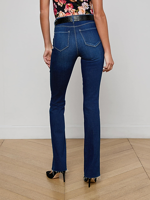 A person wearing L'AGENCE Ruth Straight-Leg Jeans stands on a light wooden floor facing away, showcasing the jeans' raw hem, paired with black heels and a floral top.