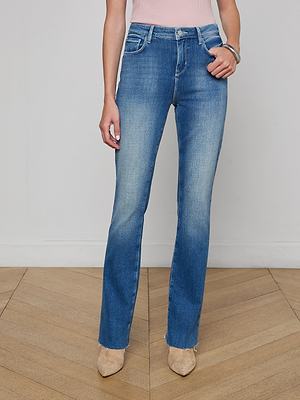 A woman models the Ruth Straight-Leg Jean, featuring a high-waisted fit and vintage wash, paired with beige pointed-toe shoes. She stands on a wooden floor against a plain white wall, with only her torso and legs visible.