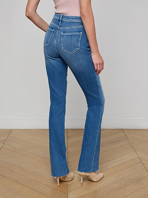 A person stands on a wooden floor facing away, wearing Ruth Straight-Leg Jeans with a raw hem and vintage wash, a light pink sleeveless top, and beige heeled shoes.