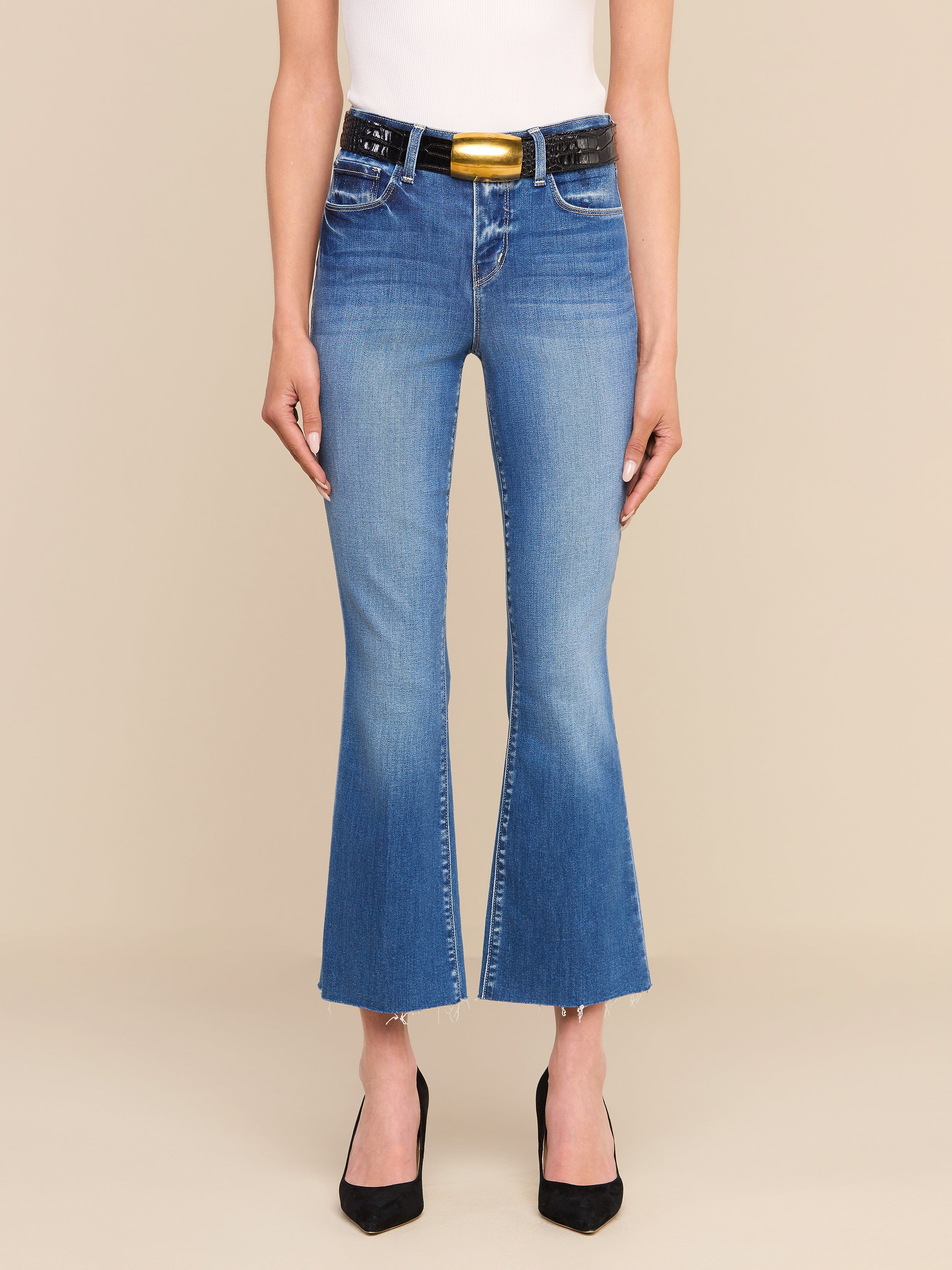 A person wearing the Kendra Cropped Flare Jean—crafted from premium stretch denim—in blue, paired with a black belt featuring a gold buckle, a white top, and black pointed heels, stands against a plain white background. Only the lower half of the body is visible.