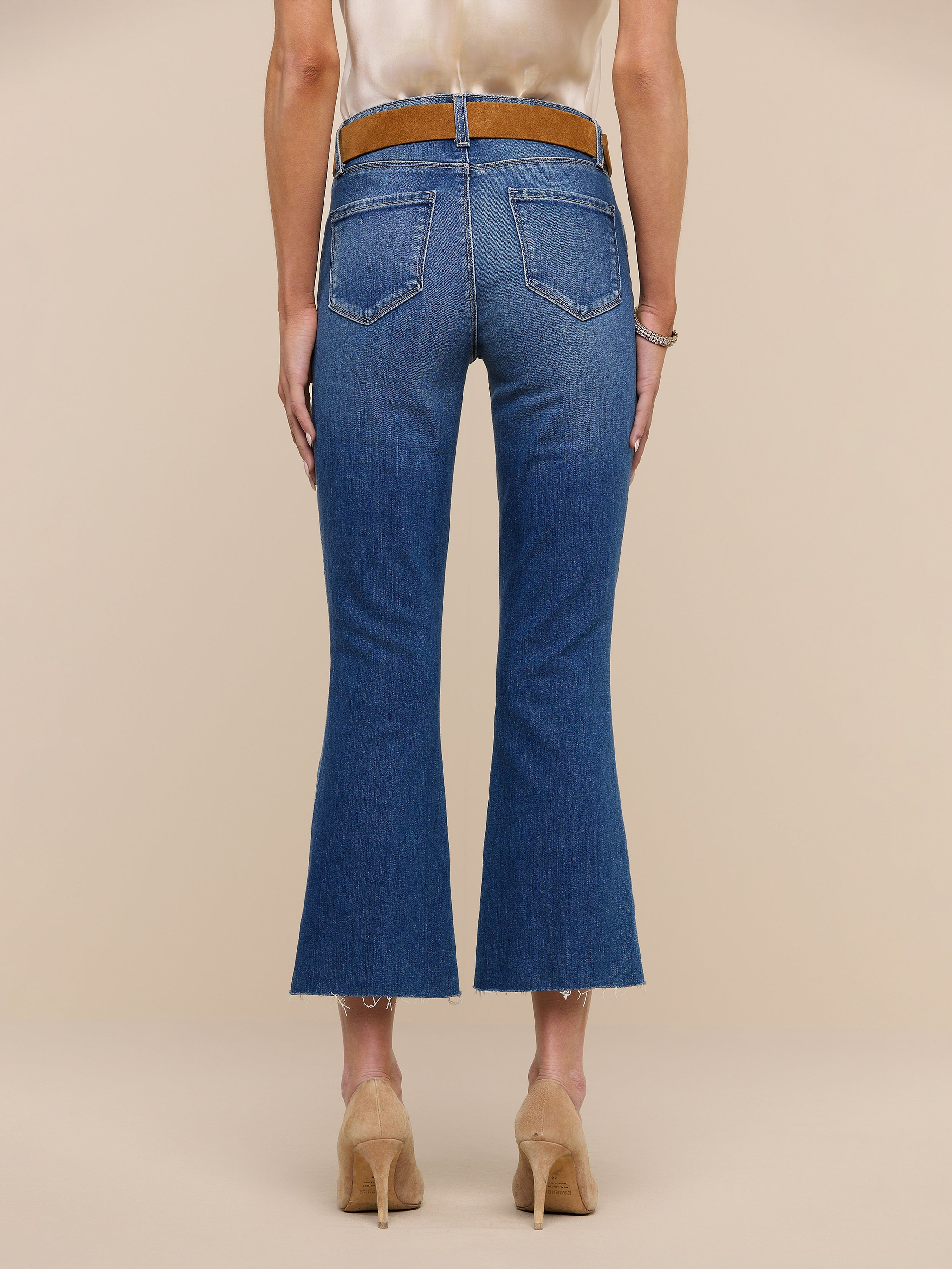 A person stands facing away on a plain white background, wearing the Kendra Cropped Flare Jean in blue—crafted from sustainable recycled fiber—paired with a white sleeveless top, a black belt, and black heeled shoes.