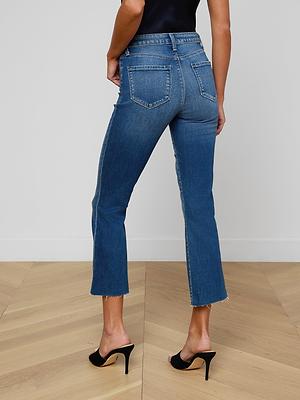 A woman stands with her back to the camera against a white wall, wearing Kendra Cropped Flare Jeans and black open-toe heels on light wood flooring.