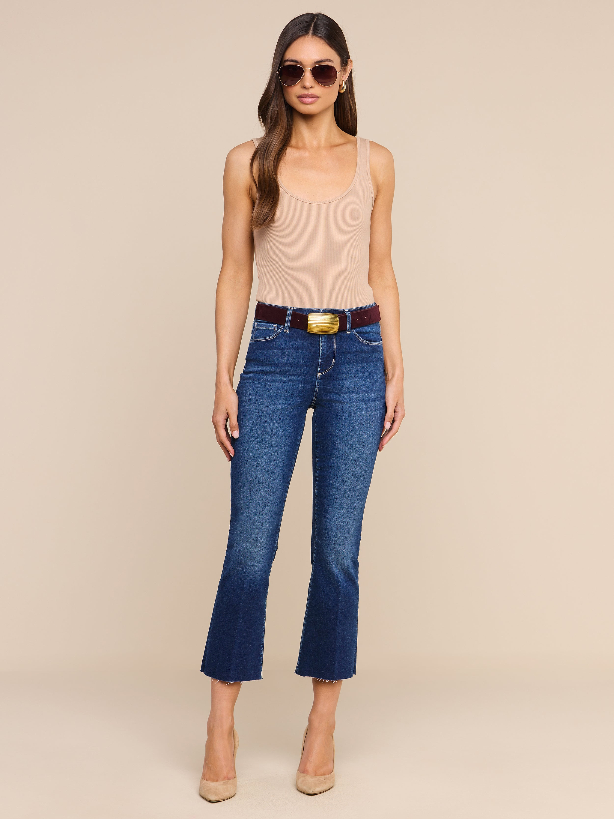 Une femme portant des lunettes de soleil, un haut sans manches beige, le jean évasé court « Kendra » avec une ceinture en denim ultra-extensible, ainsi que des talons beiges, se tient debout devant un fond beige uni.