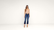 Woman wearing sunglasses, a beige sleeveless top, blue cropped jeans, and nude heels stands against a plain white background, facing forward with arms at her sides.