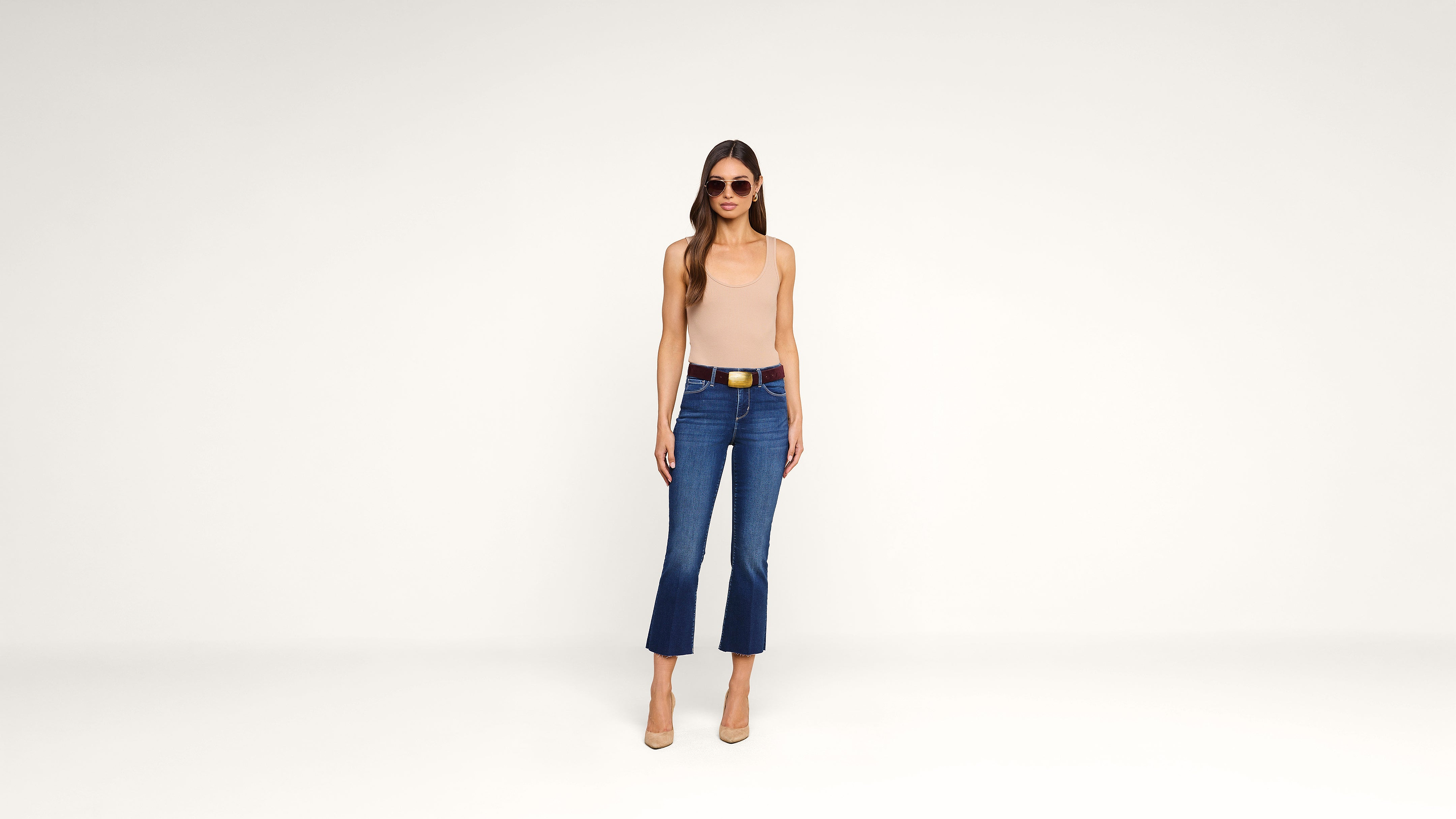 Woman wearing sunglasses, a beige sleeveless top, blue cropped jeans, and nude heels stands against a plain white background, facing forward with arms at her sides.