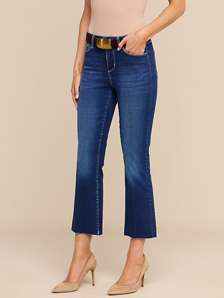 A woman stands against a plain beige background wearing the Kendra Cropped Flare Jean in blue high rise super stretch denim with a brown belt, paired with a beige sleeveless top and tan high-heeled shoes.