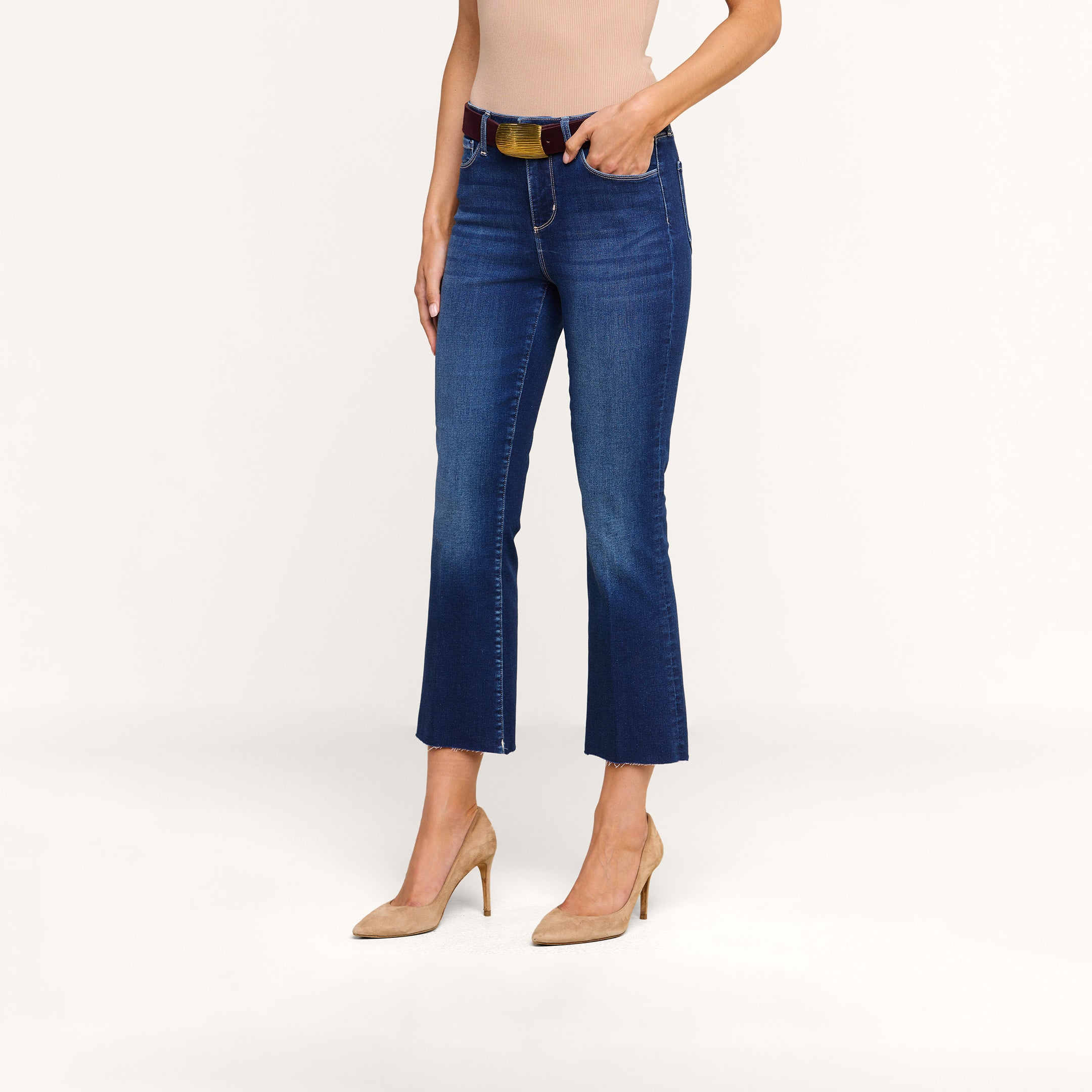 Une femme se tient debout devant un fond beige uni. Elle porte le jean « Kendra Cropped Flare » en denim bleu à taille haute ultra-extensible, agrémenté dune ceinture marron, associé à un haut sans manches beige et à des chaussures à talons hauts couleur beige clair.