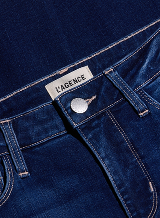 Close-up of the waistband and button of the Kendra Cropped Flare Jean in blue denim, featuring a white L’AGENCE label above the button. The visible stitching highlights the texture and details of the super stretch denim fabric.