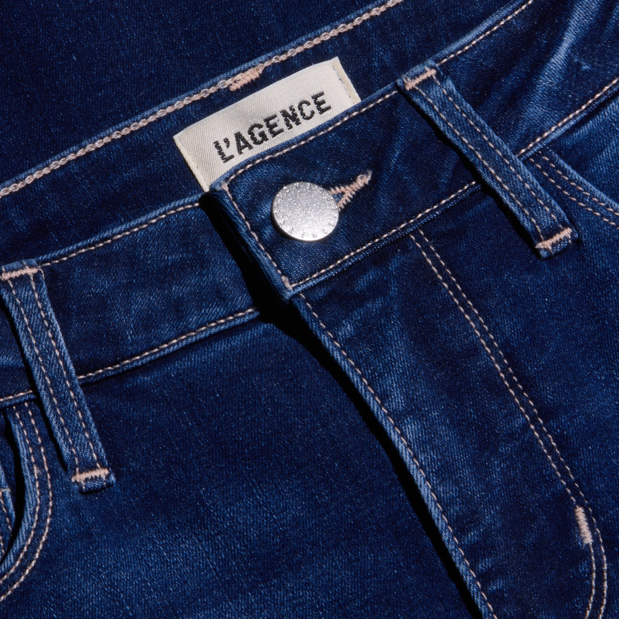 Close-up of the Kendra Cropped Flare Jean in dark blue, featuring a high rise, silver button, front pocket, visible stitching, and a white LAGENCE fabric label above the waistband. Made from super stretch denim for a comfortable fit.