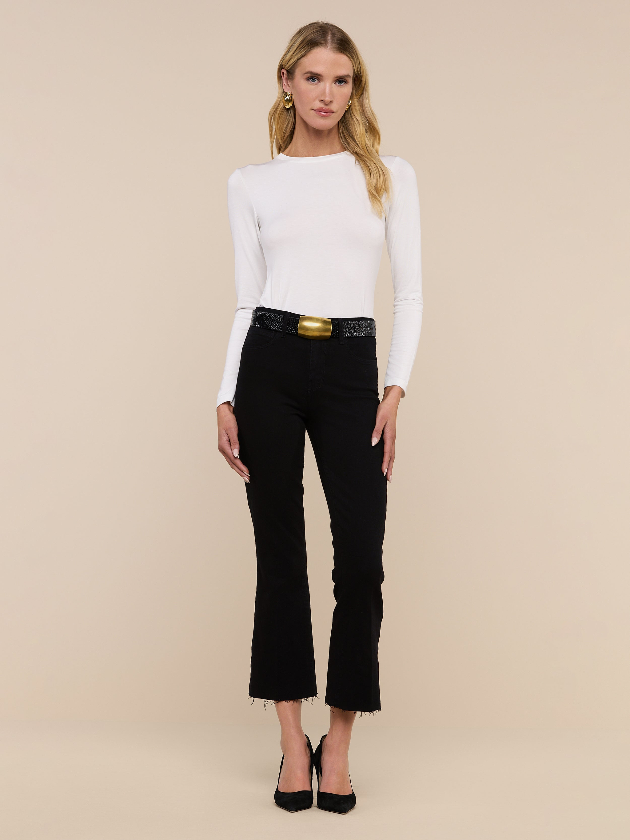 A woman stands facing forward, wearing a white long-sleeve top and the Kendra Cropped Flare Jean—premium high-rise cropped flare jeans in a black hue with frayed hems—paired with a black belt featuring a gold buckle and black heels, against a plain white background.