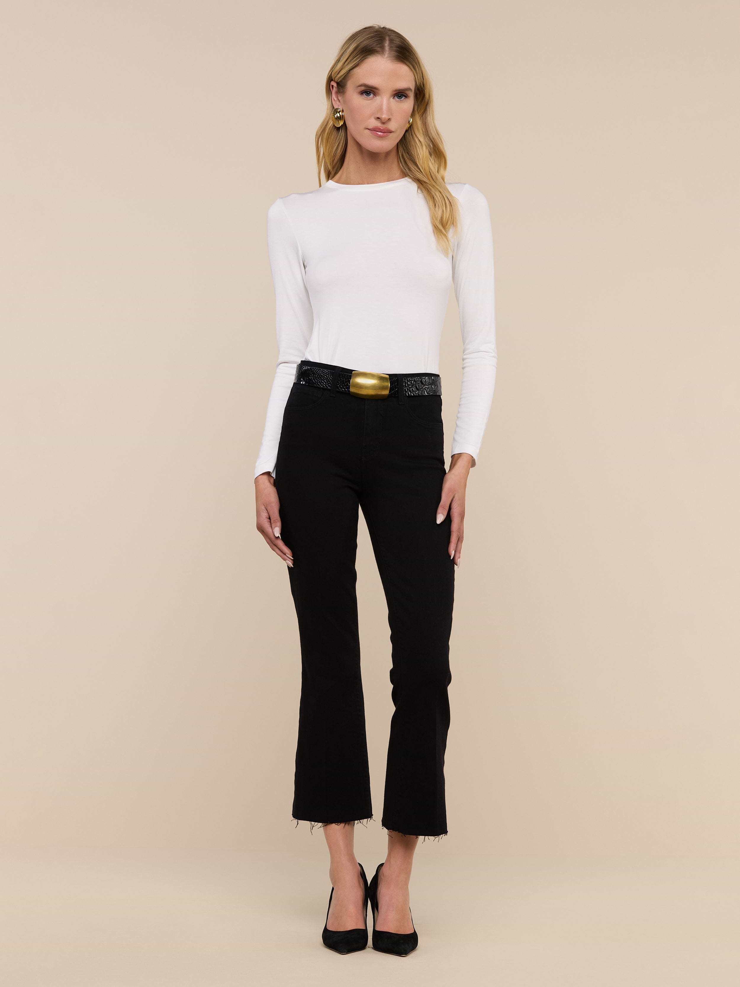 A woman stands facing forward, wearing a white long-sleeve top and the Kendra Cropped Flare Jean—premium high-rise cropped flare jeans in a black hue with frayed hems—paired with a black belt featuring a gold buckle and black heels, against a plain white background.