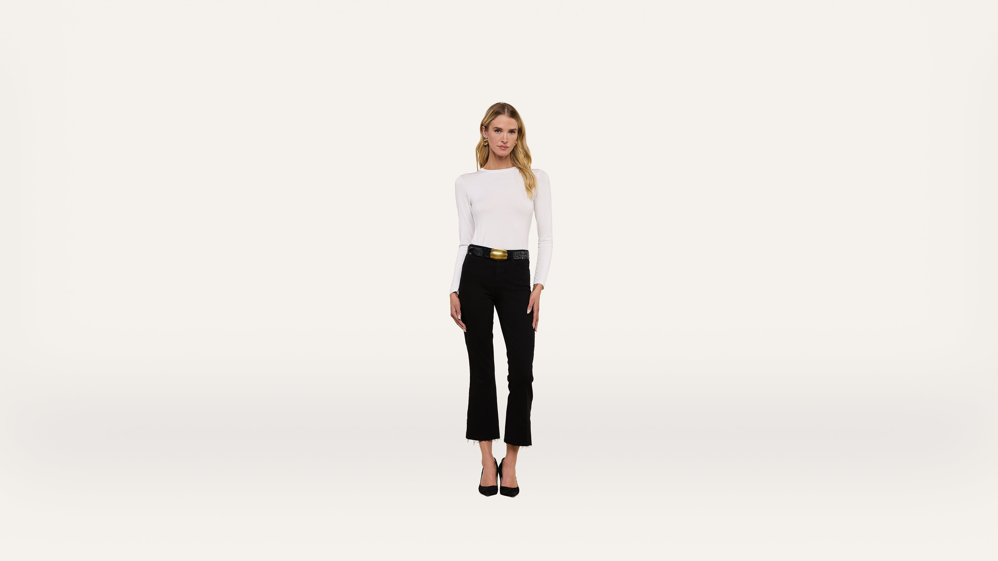 A woman stands facing forward against a plain white background, wearing a white long-sleeve top, black pants with a gold-buckled belt, and black heels.