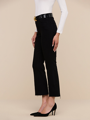 A person wears a long-sleeve white top, the Kendra Cropped Flare Jean in black premium denim, a black belt, and black high heels while standing against a beige background.