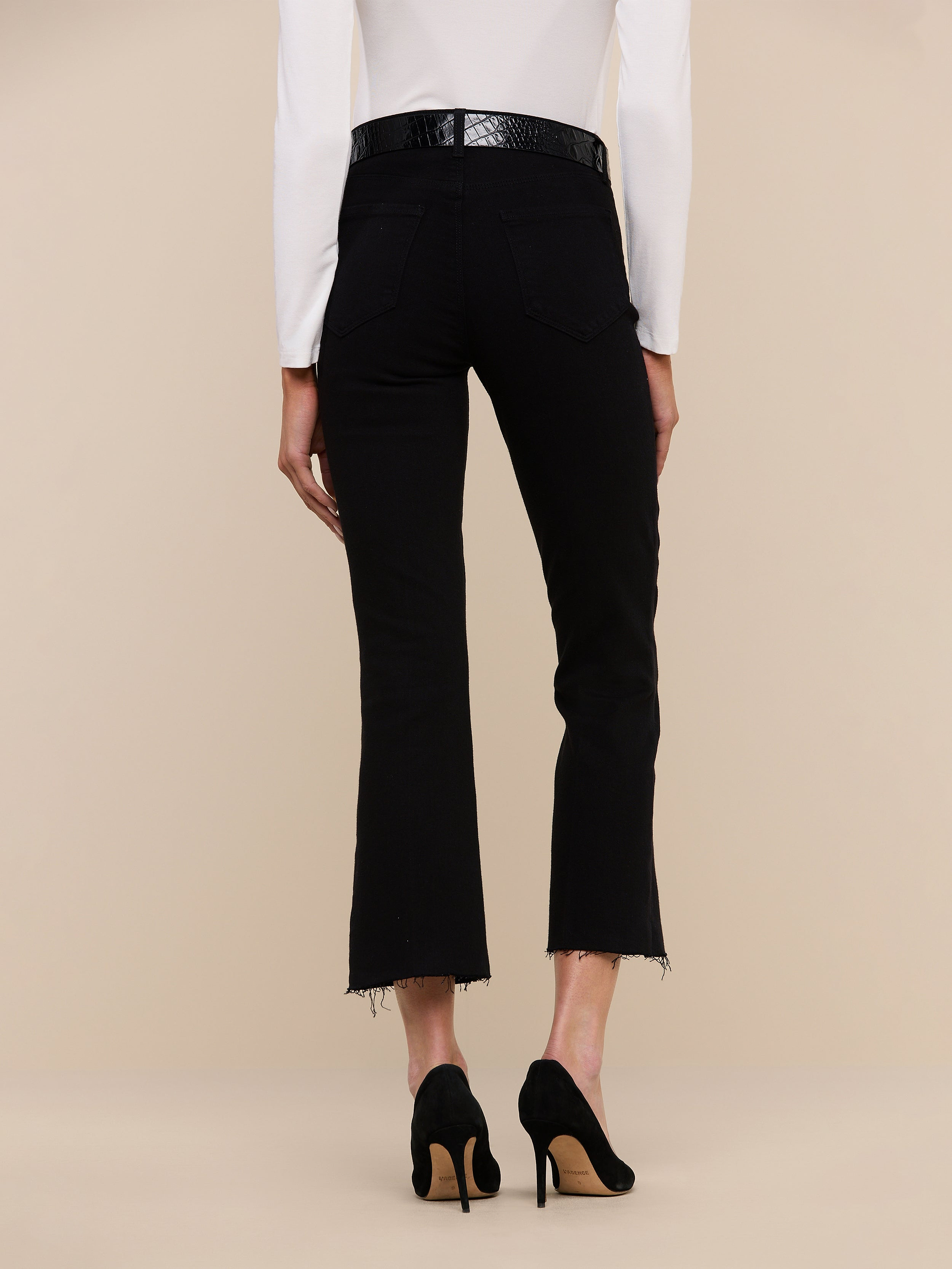 Model shown from the back against a plain light background wearing the Kendra Cropped Flare Jean in black with frayed hems, paired with black high heels and a white long-sleeve top.