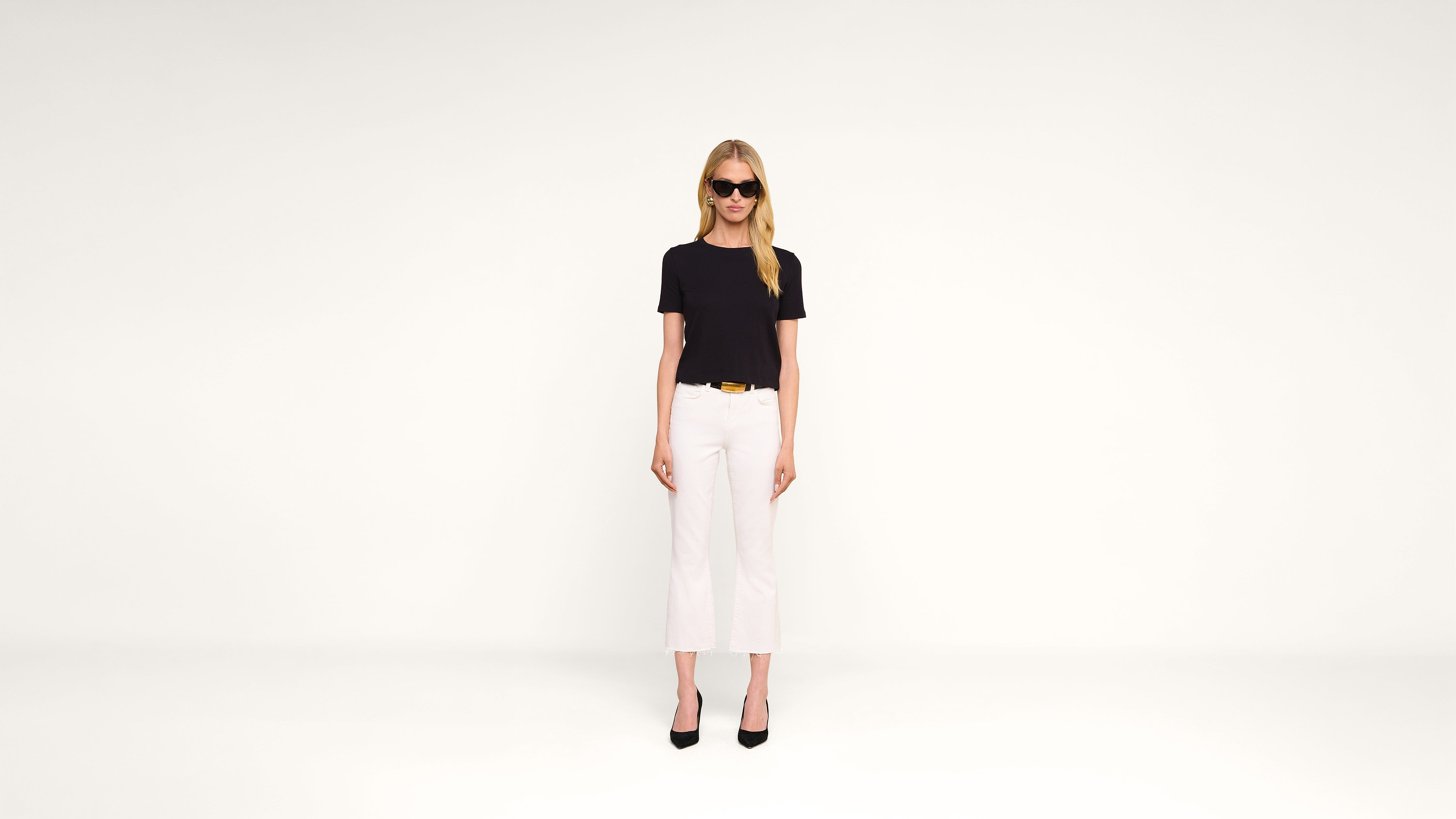 A woman wearing sunglasses, a black t-shirt, white pants, and black heels stands against a plain white background, facing forward with her arms at her sides.