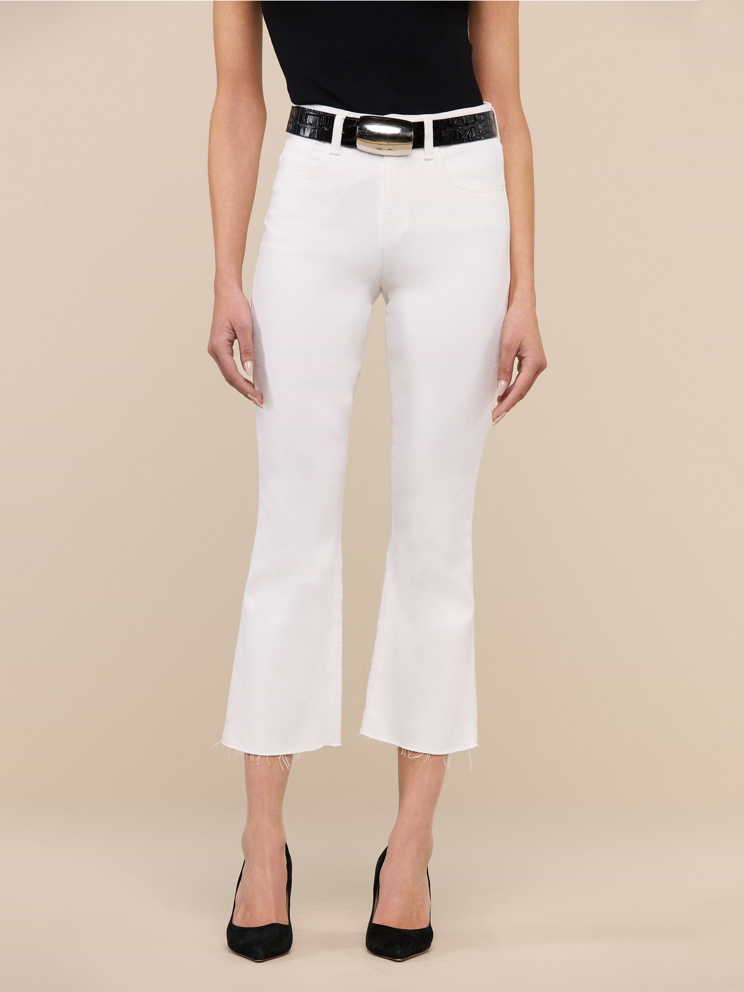 A person wearing the Kendra Cropped Flare Jean in white stretch denim, featuring a high-rise fit, cropped flare leg, and raw hem, paired with a black top and black heels stands against a plain white background. Only the lower half of the body is visible.