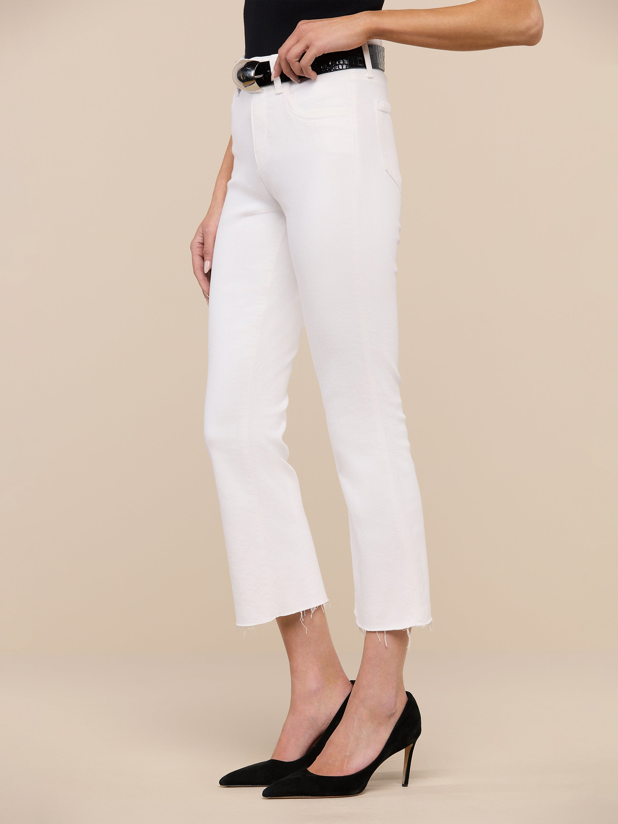 A person wearing the Kendra Cropped Flare Jean in white stretch denim with a raw hem, paired with a black top, a black belt featuring a gold buckle, and black pointed-toe heels stands against a plain white background.