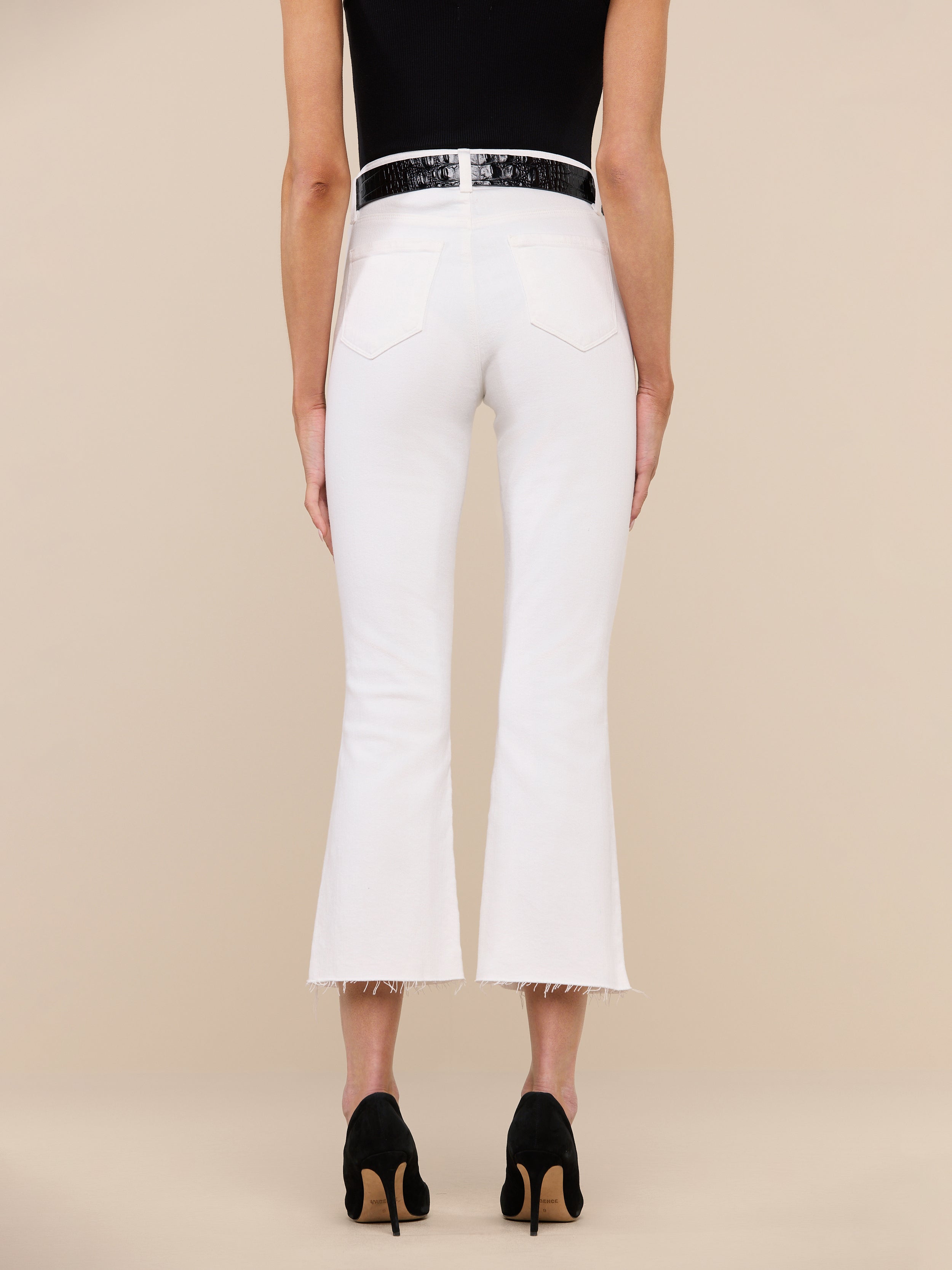 A person is standing facing away, wearing a black top, the Kendra Cropped Flare Jean in white stretch denim featuring a high-rise fit and raw hem, paired with black high-heeled shoes, against a plain white background.