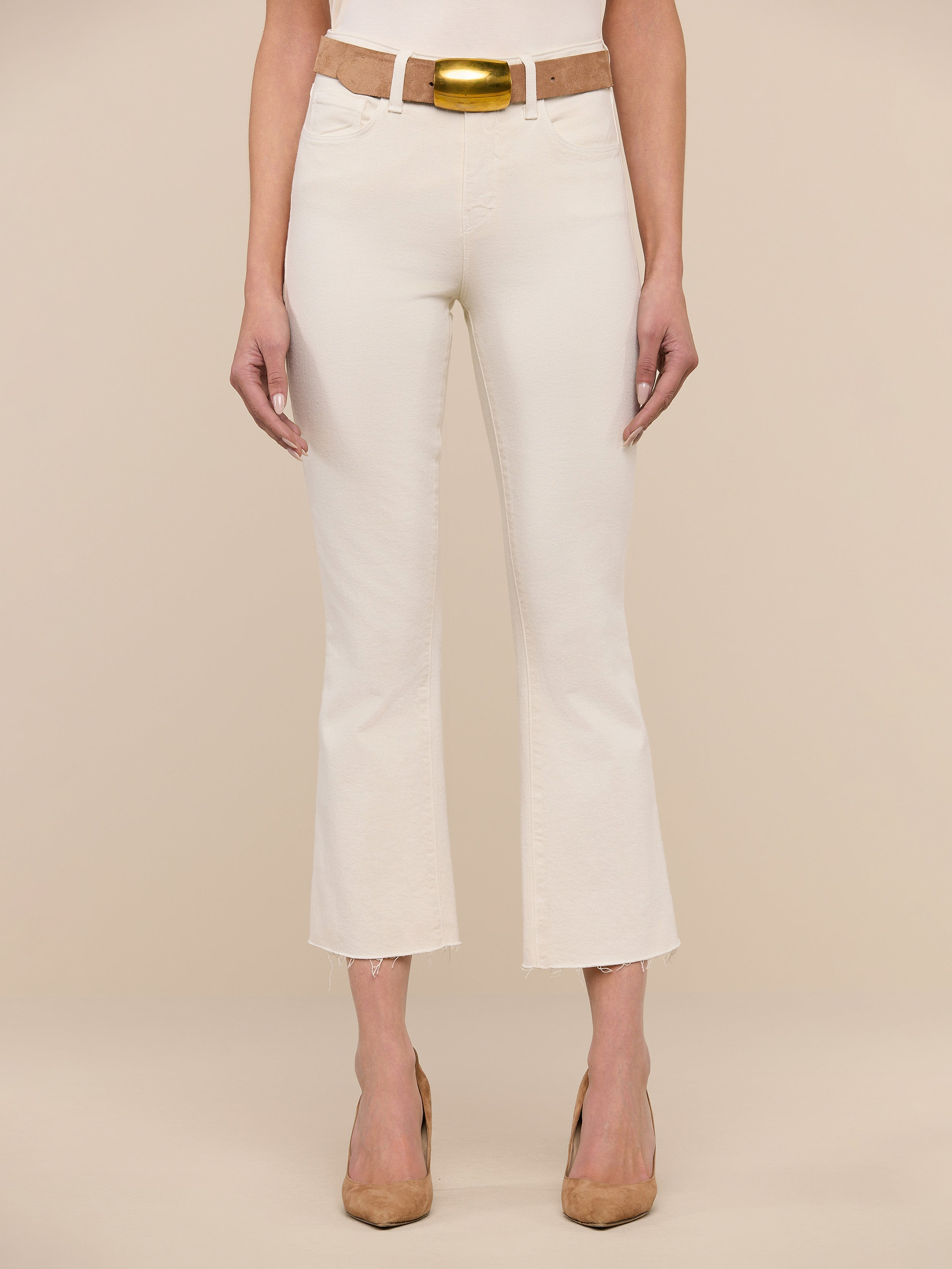 A woman stands facing forward, wearing the Kendra Cropped Flare Jean in vintage white, paired with a tan belt featuring a gold buckle and tan heels. The background is plain white.