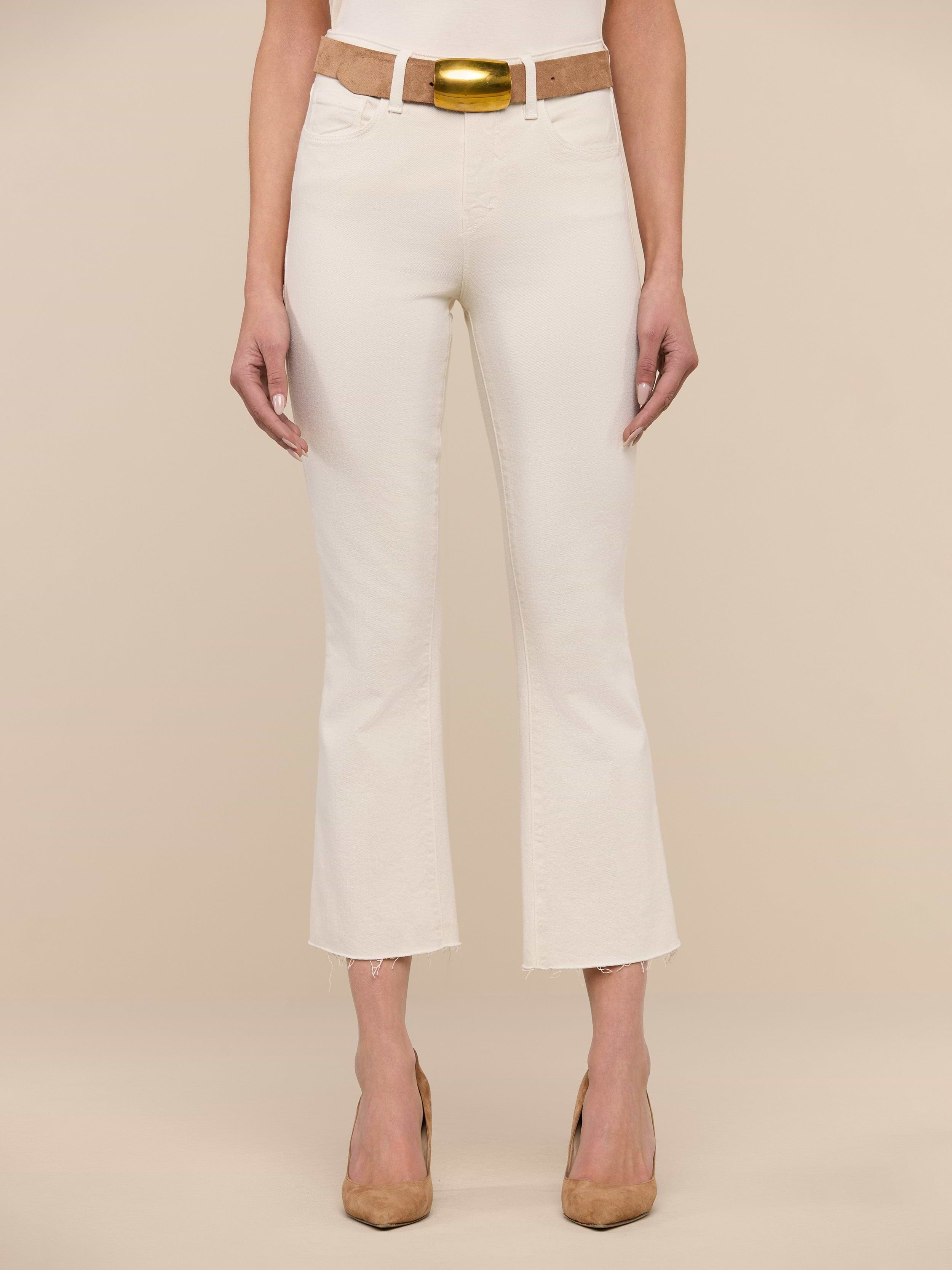 A woman stands facing forward, wearing the Kendra Cropped Flare Jean in vintage white, paired with a tan belt featuring a gold buckle and tan heels. The background is plain white.