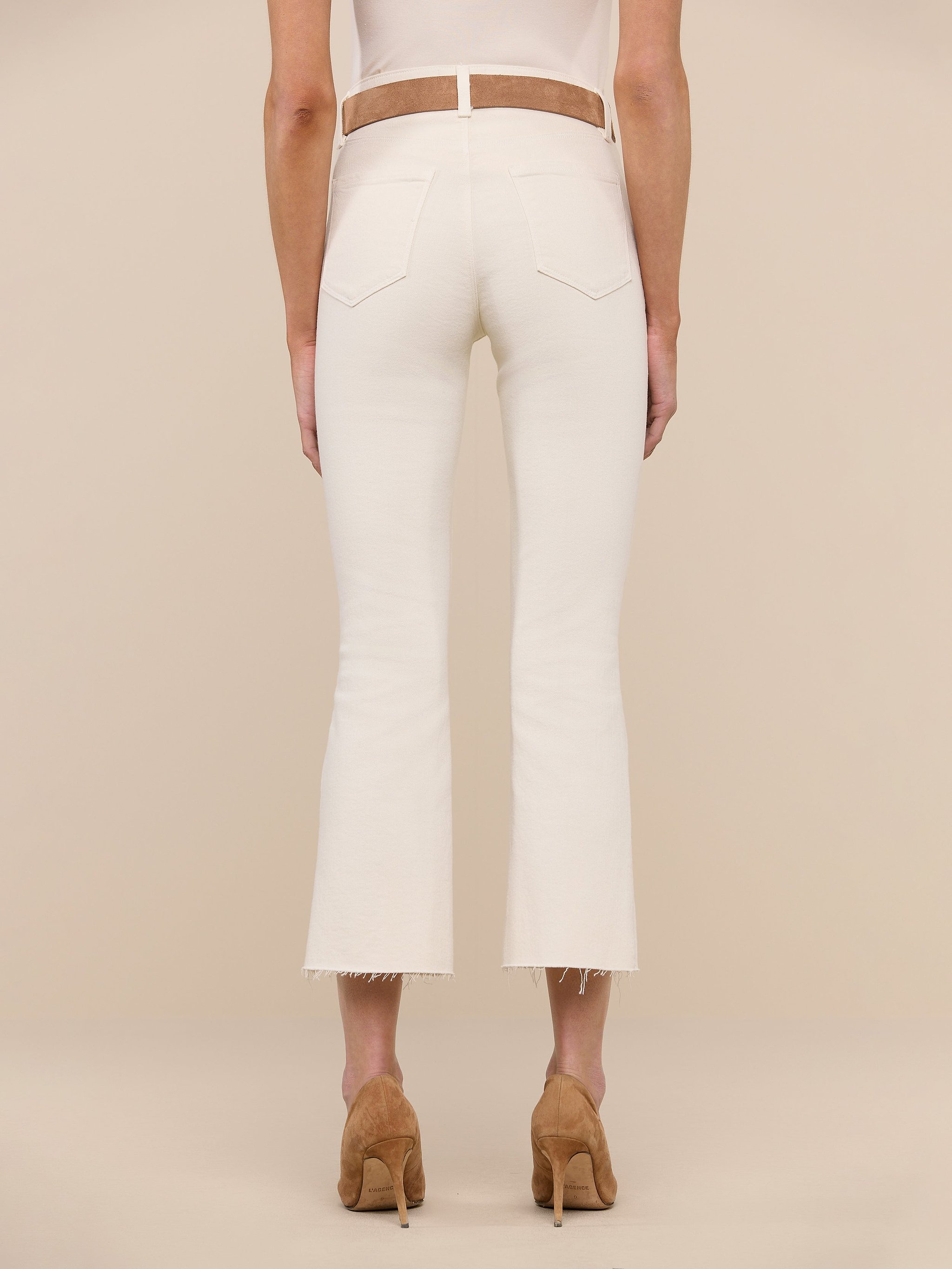 A person stands on a white background, wearing the Kendra Cropped Flare Jean in a vintage-white wash styled with a brown belt and tan high heels. The view is from behind, highlighting the back pockets and frayed hems of the jeans.