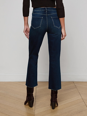 A person stands with their back to the camera, wearing L'AGENCE Kendra Cropped Flare Jean in dark wash denim, a black long-sleeve top, and dark heeled boots on a light wooden floor against a plain white wall.