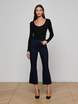A woman stands indoors against a plain light wall, wearing a fitted black long-sleeve top, L’AGENCE Kendra Cropped Flare Jeans in super stretch indigo denim, and black heels, with one hand in her pocket.