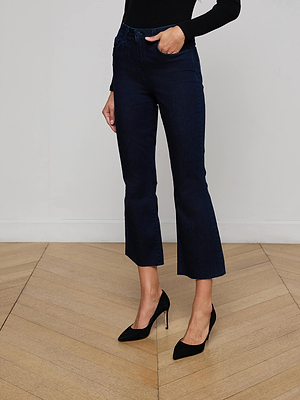 A woman stands on a wooden floor in L’AGENCE’s Kendra Cropped Flare Jean in dark indigo super stretch denim, paired with a black long-sleeve top and black pointed high heels. Her torso is out of frame, one hand on her hip.