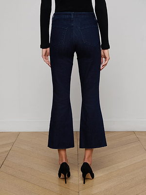 A person wearing L'AGENCE Kendra Cropped Flare Jean in dark indigo wash with black heels and a long-sleeve black top stands on a wooden floor, facing away from the camera.