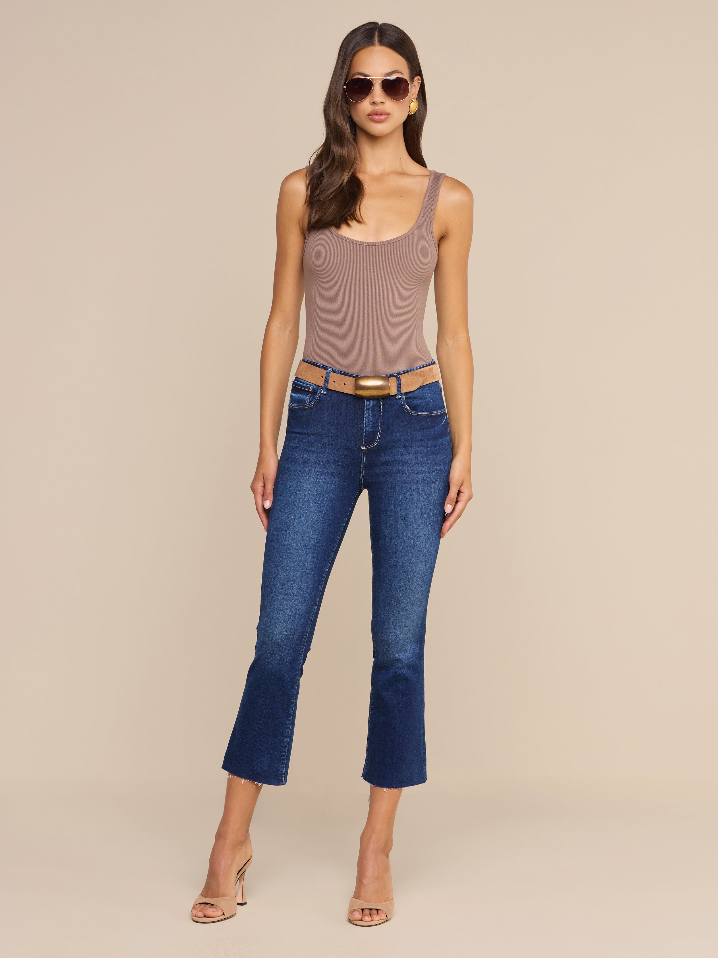 A woman wearing sunglasses, a sleeveless taupe bodysuit, heeled sandals, and the Kendra Cropped Flare Jean stands against a plain white background.