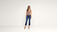 A woman wearing sunglasses, a beige sleeveless top, blue cropped jeans, and heels stands against a plain white background, facing forward with her arms at her sides.