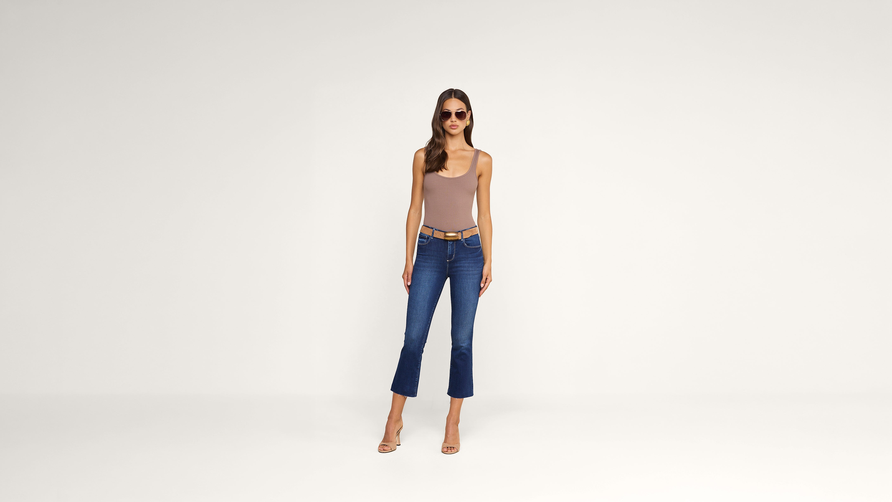 A woman wearing sunglasses, a beige sleeveless top, blue cropped jeans, and heels stands against a plain white background, facing forward with her arms at her sides.
