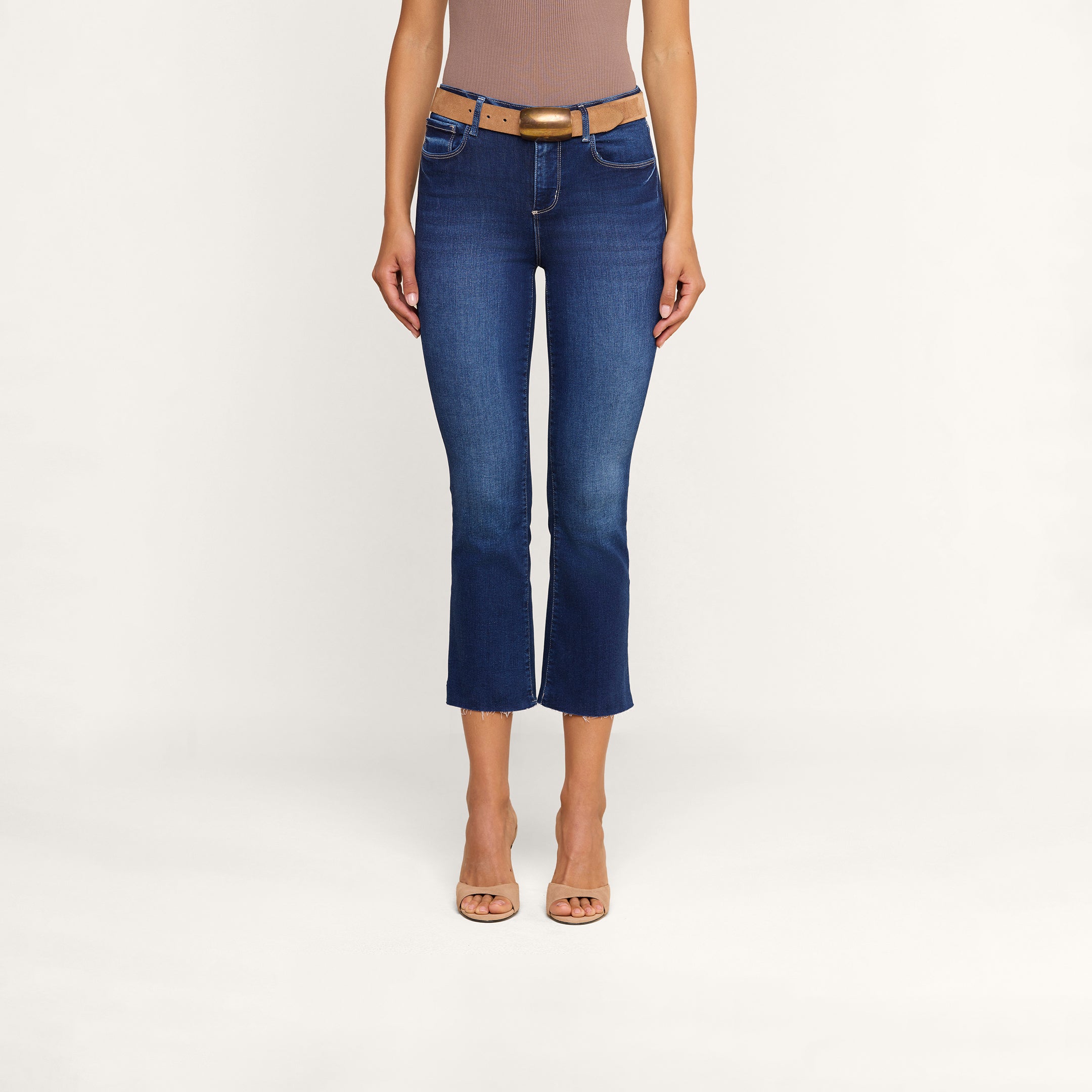 A woman stands against a plain white background wearing a sleeveless top, open-toed heels, a tan belt, and the Kendra Cropped Flare Jean—blue jeans with a raw hem made from sustainable recycled fiber.