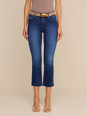 A woman stands against a plain beige background wearing Kendra Cropped Flare Jeans with raw hems, a tan belt, and beige open-toe heels.