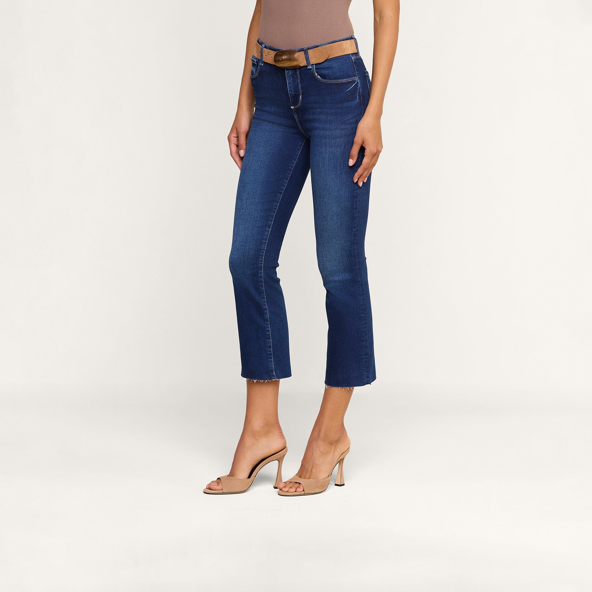 Wearing the Kendra Cropped Flare Jean—crafted from sustainable recycled fiber—a woman pairs her blue cropped flare jeans with a brown sleeveless top, a brown belt, and beige high-heeled shoes as she stands against a plain white background.
