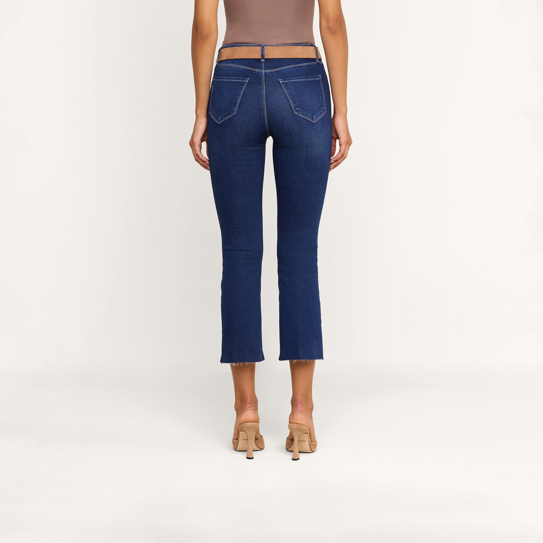 A person wearing high-heeled shoes and the Kendra Cropped Flare Jean, made from stretch denim with a sheer waistband and back pockets, is seen from behind against a plain white background.