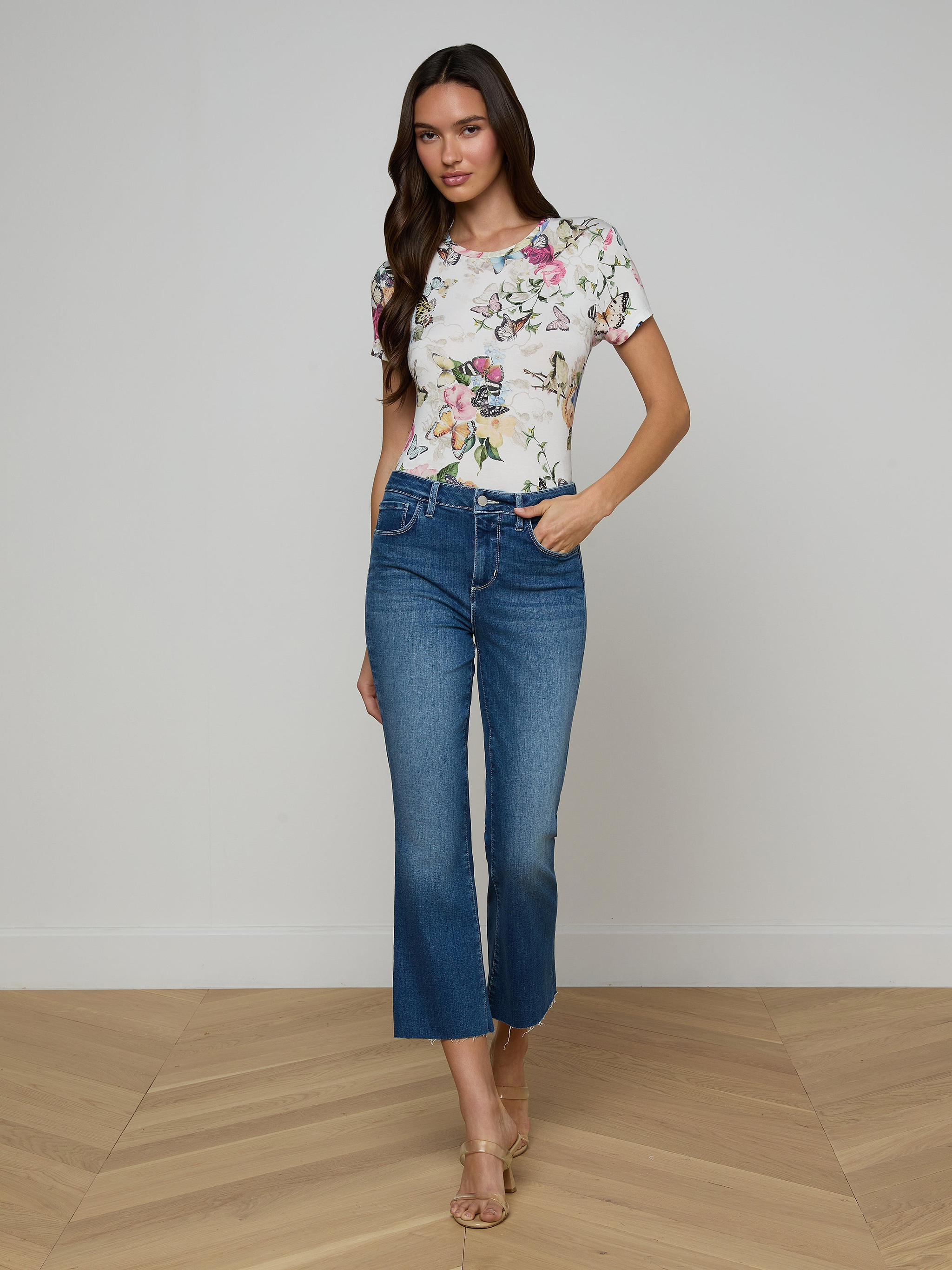 A woman stands against a plain white background, wearing a floral short-sleeve top, beige heeled sandals, and the Kendra Cropped Flare Jean—a pair of blue high-waisted cropped flare jeans in a dark denim wash. She has long brown hair and one hand in her pocket.