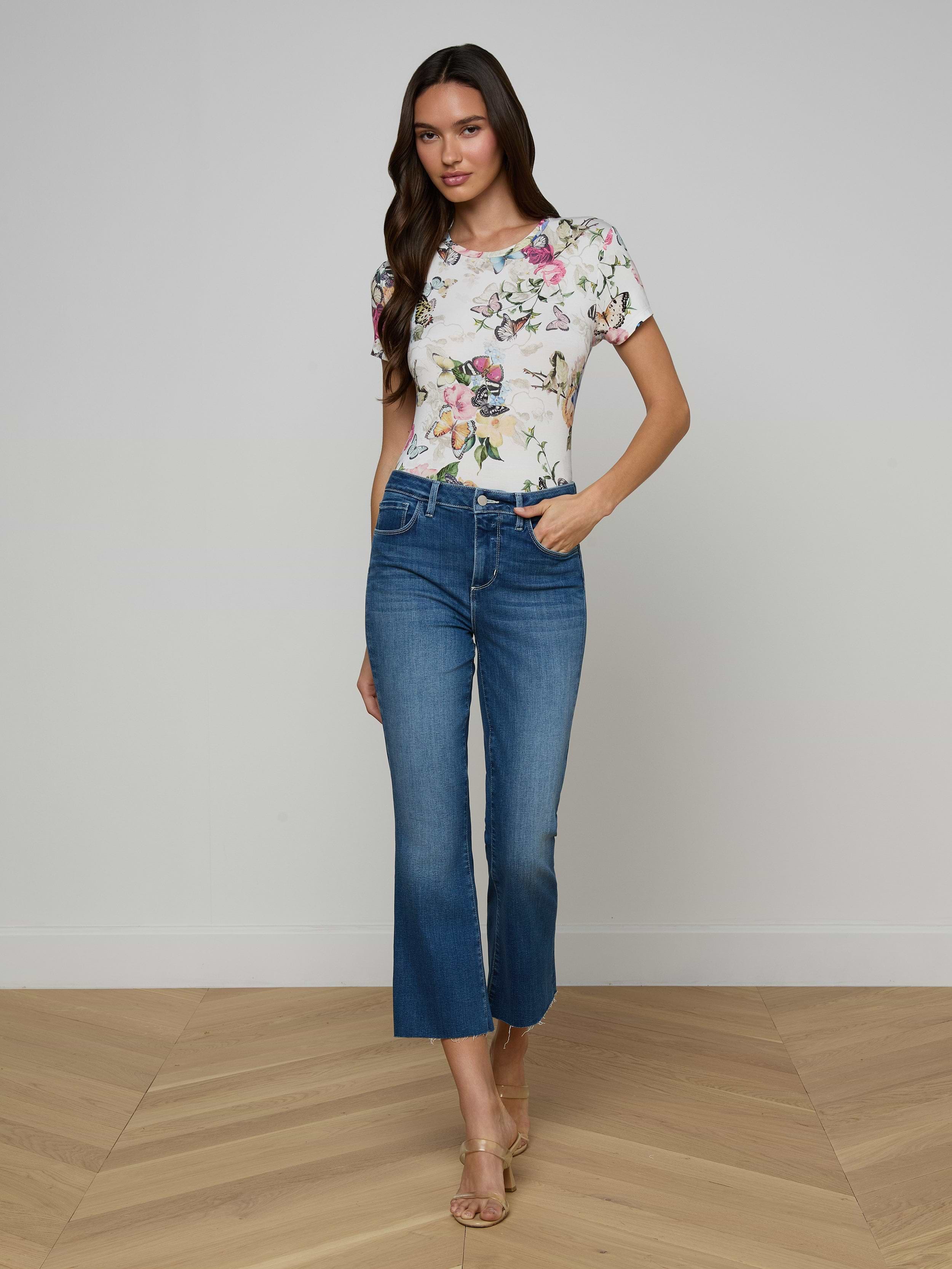 A woman stands against a plain white background, wearing a floral short-sleeve top, beige heeled sandals, and the Kendra Cropped Flare Jean—a pair of blue high-waisted cropped flare jeans in a dark denim wash. She has long brown hair and one hand in her pocket.