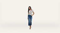 A woman stands against a plain white background, wearing a floral short-sleeve top, blue cropped jeans, and nude heeled sandals.