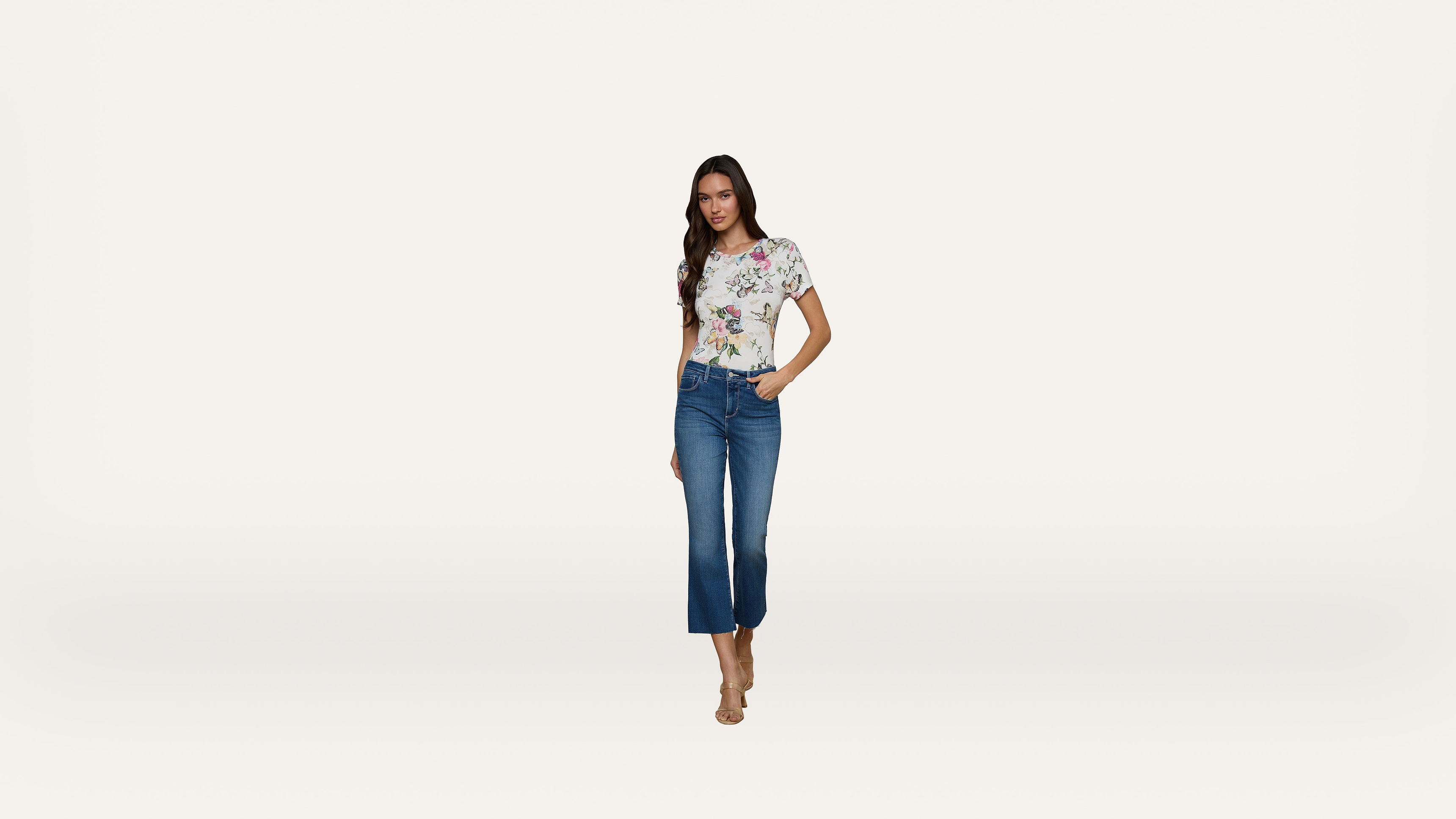 A woman stands against a plain white background, wearing a floral short-sleeve top, blue cropped jeans, and nude heeled sandals.