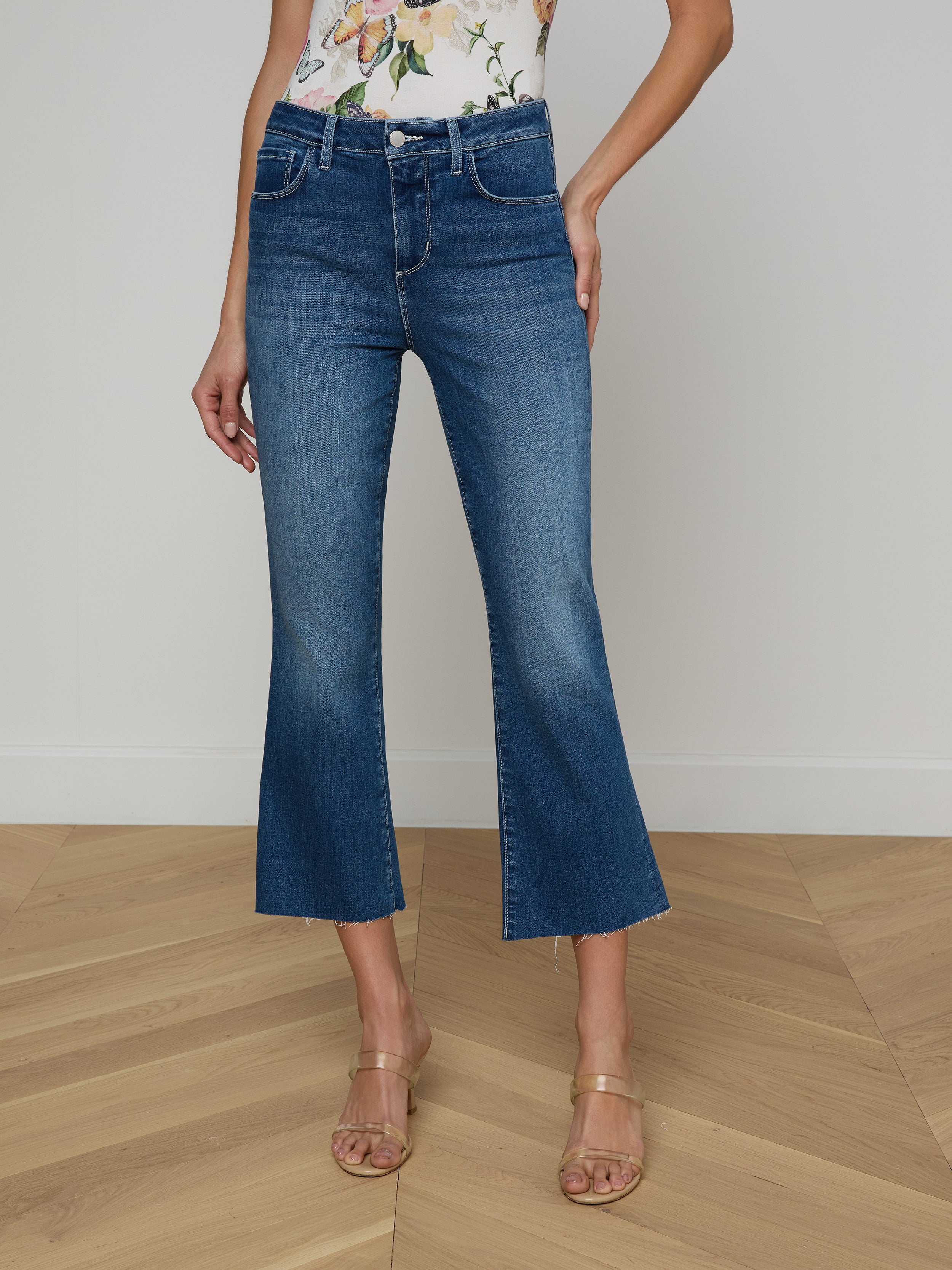 Wearing the Kendra Cropped Flare Jean in a dark denim wash, a person pairs it with a floral sleeveless top and beige heeled sandals while standing against a plain white background. Only the lower half of their body is visible.