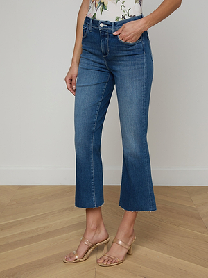 Someone wears the Kendra Cropped Flare Jean in super stretch denim with a raw hem, paired with a floral top and clear heeled sandals, standing on a light wooden floor against a plain white wall.