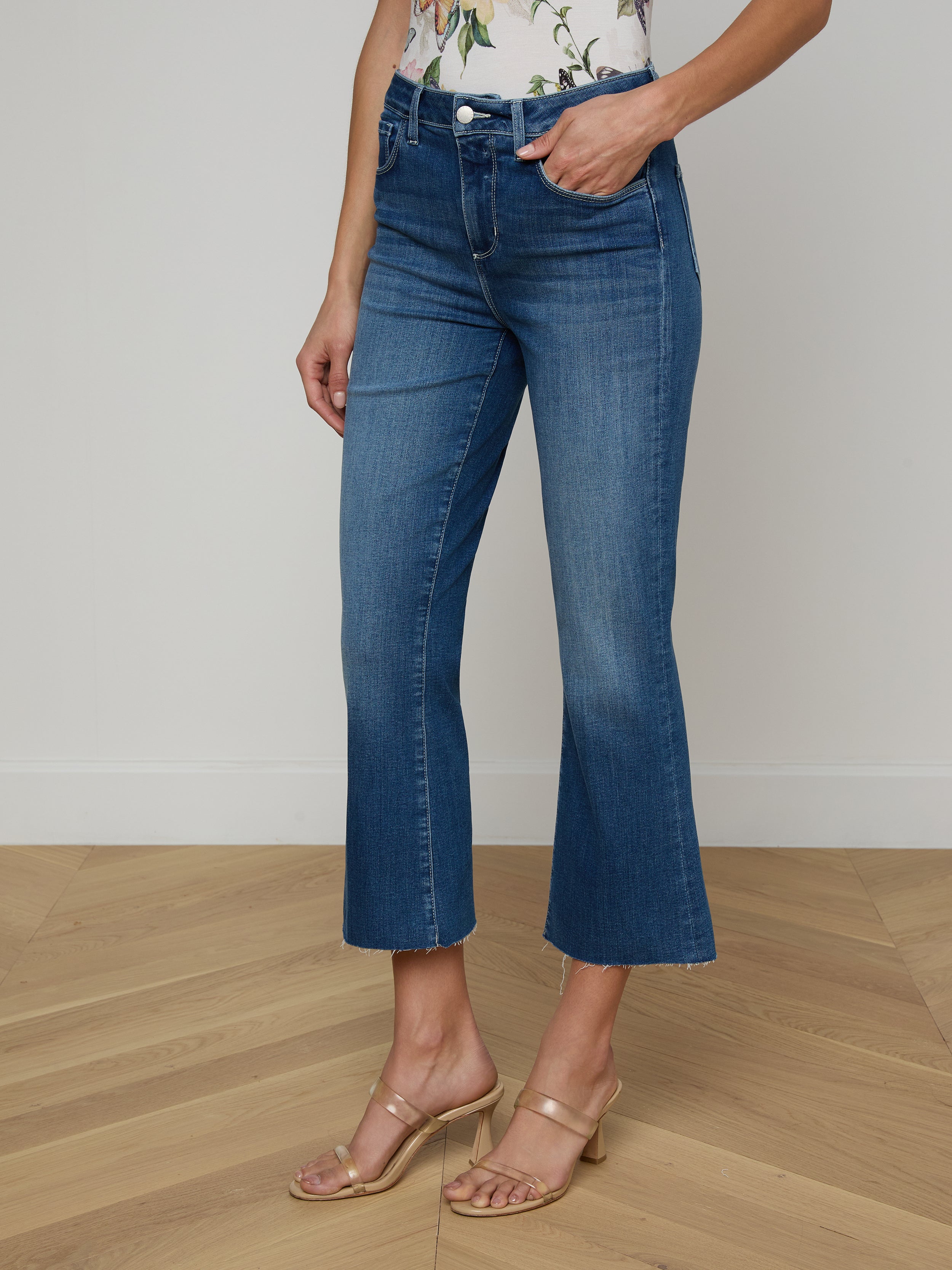 A person models the Kendra Cropped Flare Jean in a dark denim wash, paired with a floral top and beige heeled sandals, posing against a plain white background with only the lower half of the body visible.