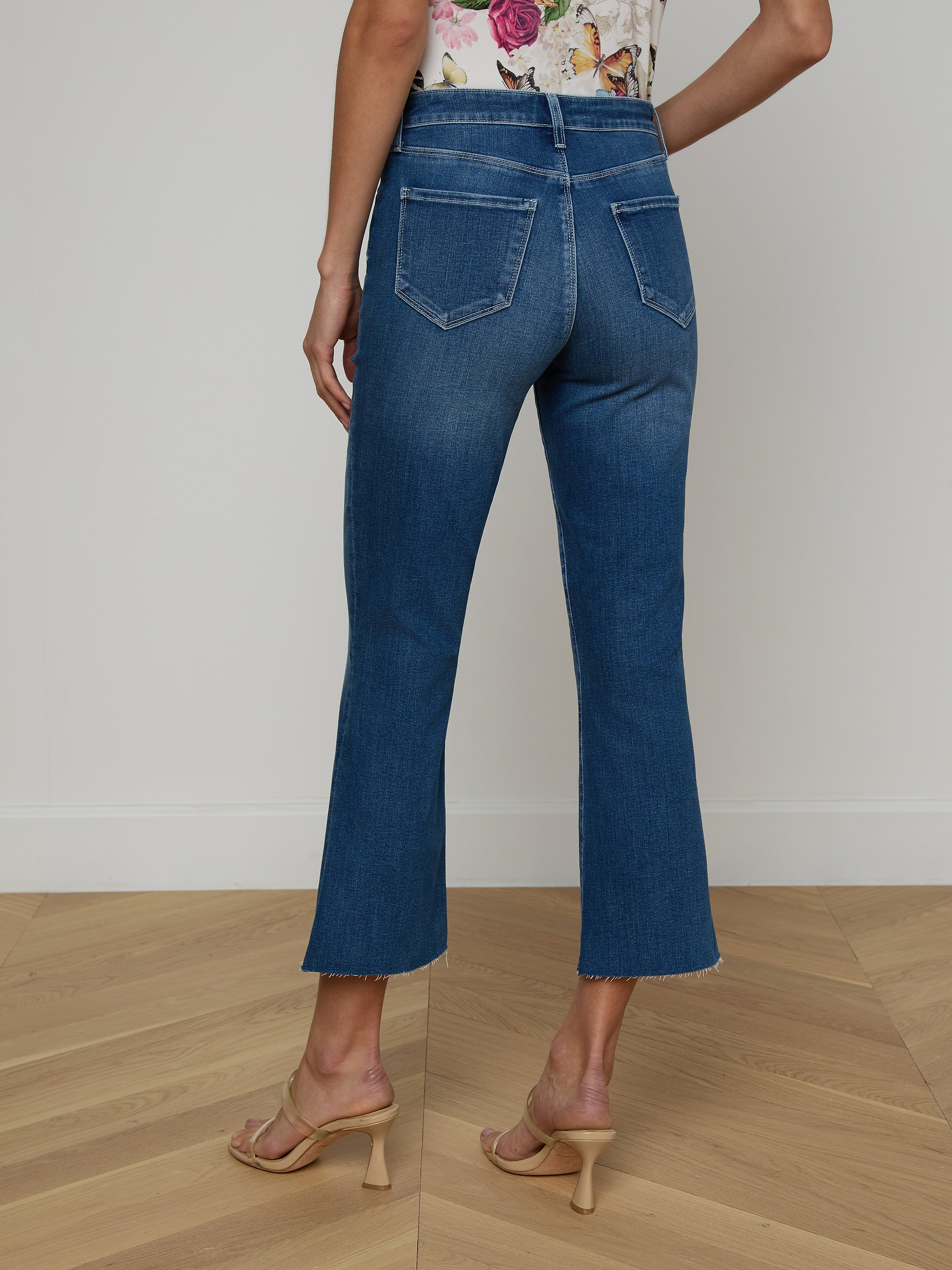 A woman stands facing away against a plain white background, showcasing the back view of the Kendra Cropped Flare Jean in a dark blue denim wash. She pairs the jeans with a floral top and tan heeled sandals.