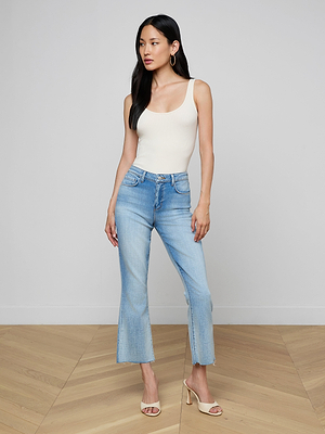 A woman with long dark hair and gold hoop earrings stands indoors on a wooden floor, wearing a cream sleeveless top, beige heeled sandals, and Kendra Cropped Flare Jeans in light wash stretch denim.