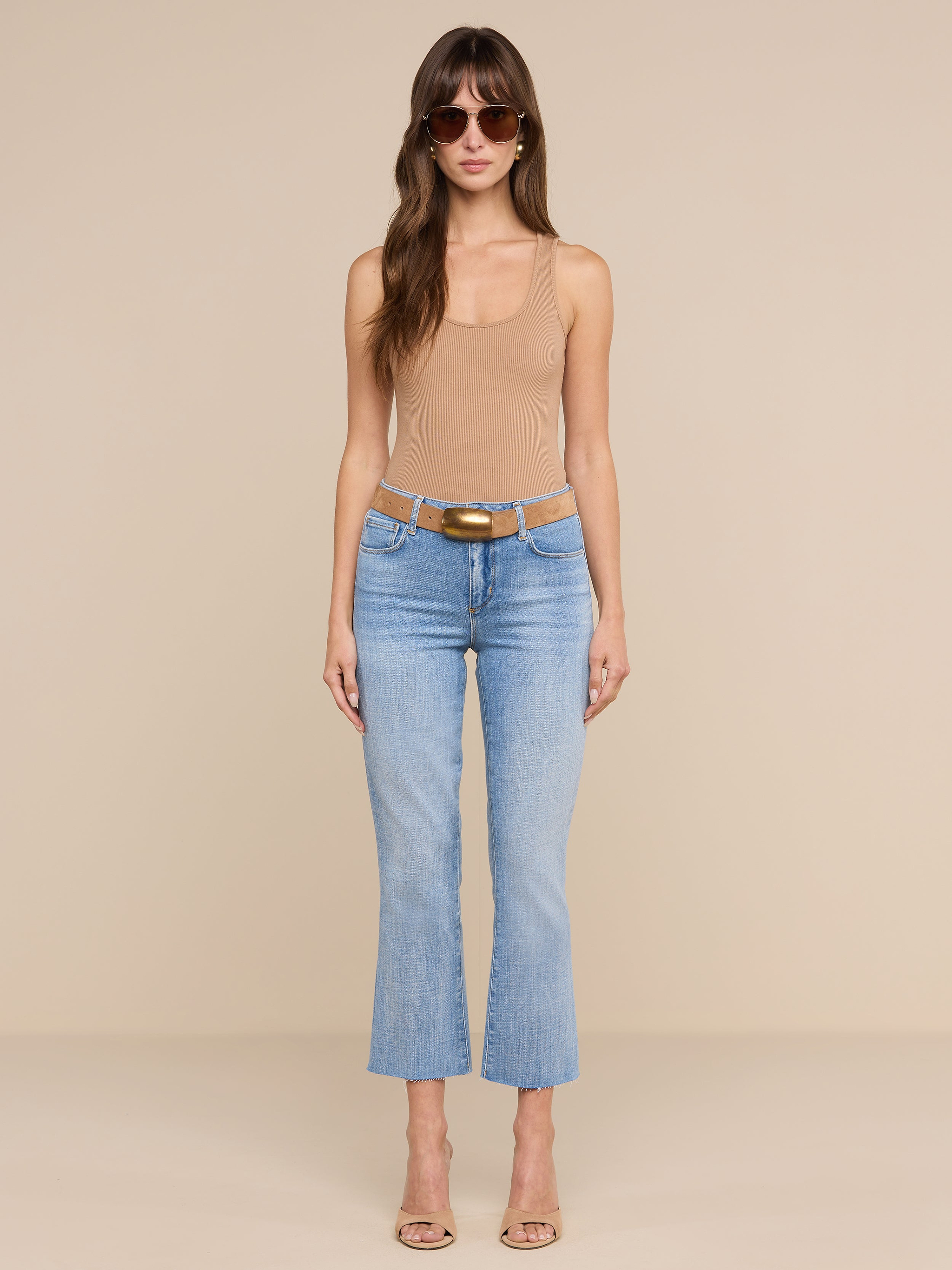 A woman wearing sunglasses, a tan sleeveless top, the Kendra Cropped Flare Jean in light wash, a brown belt, and beige heels stands facing forward against a plain white background.