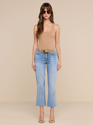 Una donna è in posa su uno sfondo beige a tinta unita e indossa i jeans Kendra Cropped Flare, occhiali da sole, una canotta beige, una cintura color cuoio e scarpe con tacco aperte in punta.