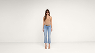 A woman wearing sunglasses, a beige sleeveless top, blue jeans, and heeled sandals stands against a plain white background.