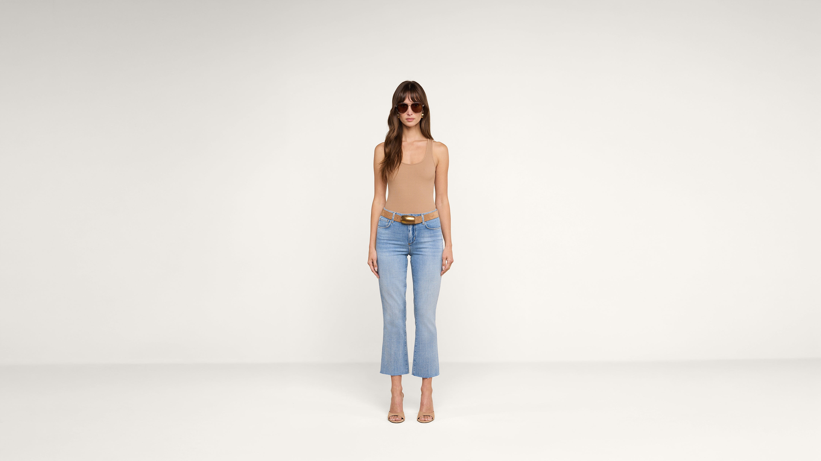 A woman wearing sunglasses, a beige sleeveless top, blue jeans, and heeled sandals stands against a plain white background.